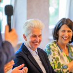 Smiling people at an event, someone speaking.